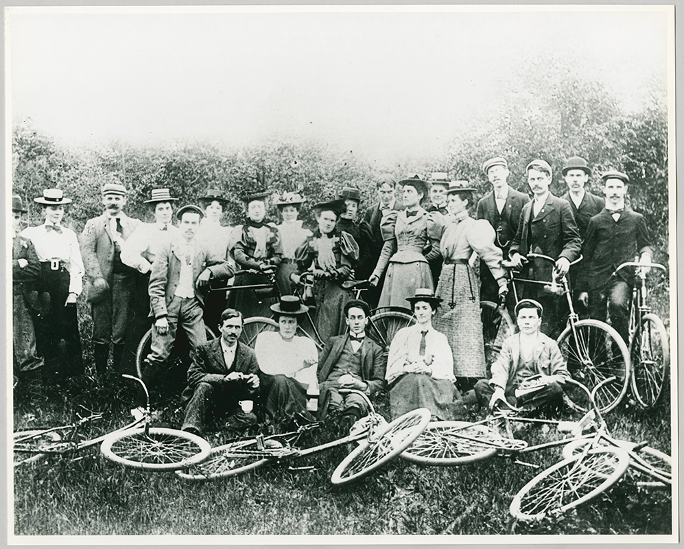 Group of adults standing and sitting together with bicycles on the ground and trees in the background.