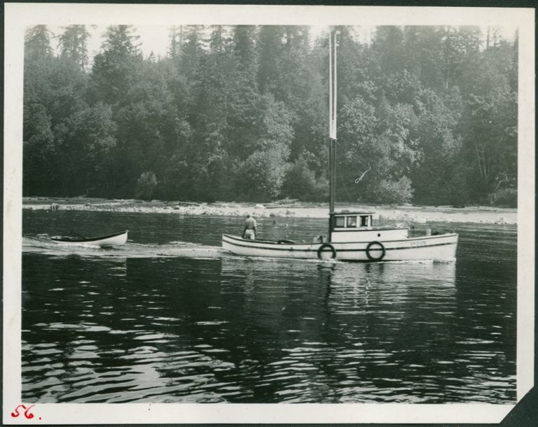 Small boat on the water with trees in the background. The boat is towing a smaller canoe like boat behind it..