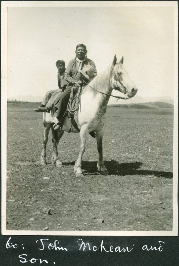 An Indigenous father and child ride a white horse on plains.