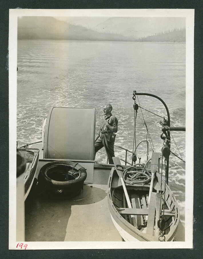An older white man stands on the front of a boat, looking towards the water.