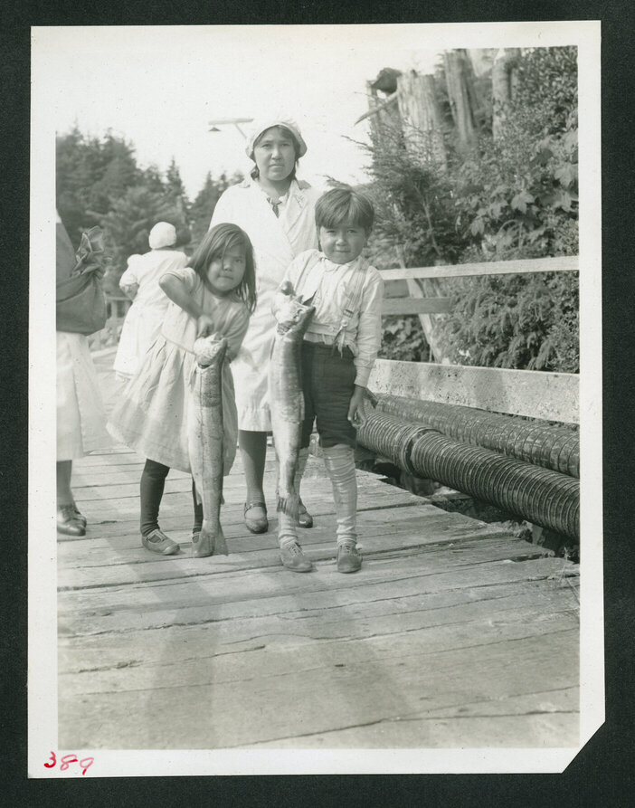 Two Indigenous children and their mother stand on a boardwalk. The two children are holding fish.